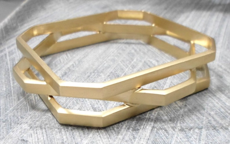 May include: Gold-colored geometric bangle bracelets. The bracelets have a unique, angular design with interlocking shapes. The metal has a matte finish, and the bracelets are displayed on a textured, gray surface. The overall aesthetic is modern and minimalist.