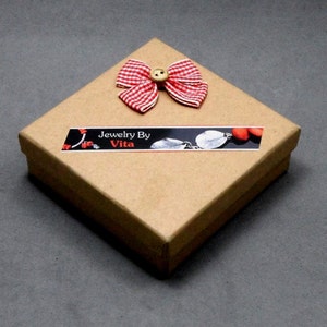 May include: A square, brown cardboard gift box with a red and white checkered bow and a small wooden button. The box has a label that reads "Jewelry By Vita". The box is closed and sits on a gray surface.