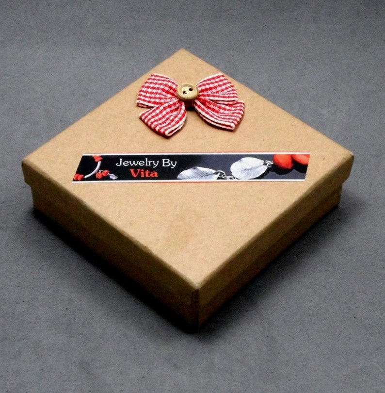 May include: A square, brown cardboard gift box with a red and white checkered bow and a small wooden button. The box has a label that reads "Jewelry By Vita". The box is closed and sits on a gray surface.