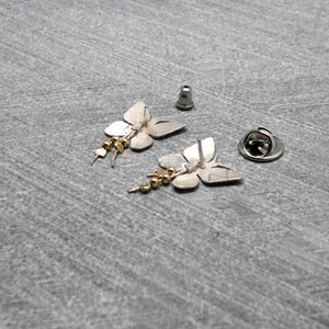 May include: A pair of silver butterfly earrings with gold beads dangling from the bottom. The earrings are shown on a gray surface with the backs of the earrings facing up.