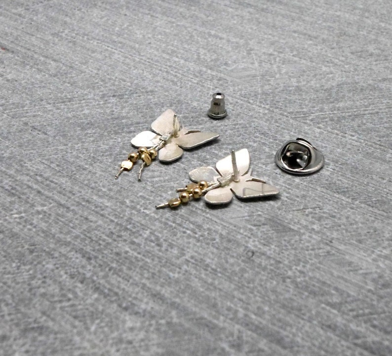 May include: A pair of silver butterfly earrings with gold beads dangling from the bottom. The earrings are shown on a gray surface with the backs of the earrings facing up.