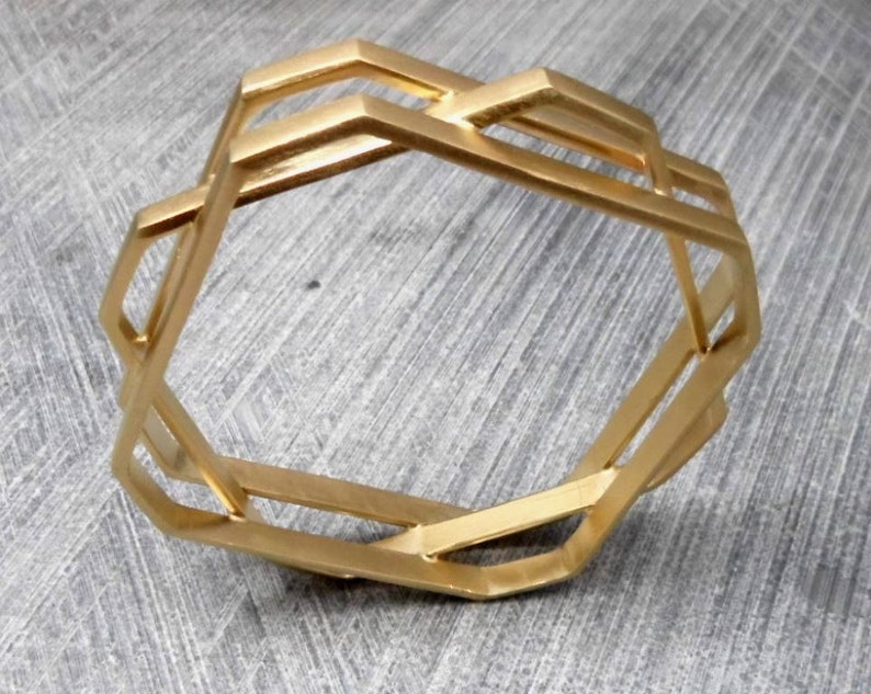 May include: A gold-colored geometric bracelet with an open design. The bracelet features a series of interconnected, angular shapes, creating a modern and stylish accessory. The bracelet is made of metal and has a matte finish.