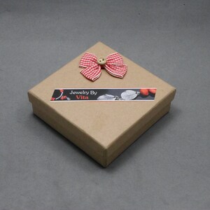 May include: A small, square, brown cardboard box with a red and white gingham bow and a button on top. The box has a label that says "Jewelry By Vita".