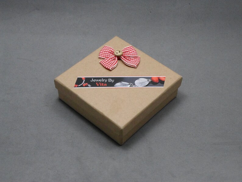 May include: A small, square, brown cardboard box with a red and white gingham bow and a button on top. The box has a label that says "Jewelry By Vita".