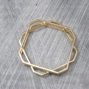 May include: A set of three gold-colored geometric bangles. Each bangle has a unique octagonal shape with interlocking designs. The bangles are displayed on a gray surface, showcasing their modern and minimalist style.