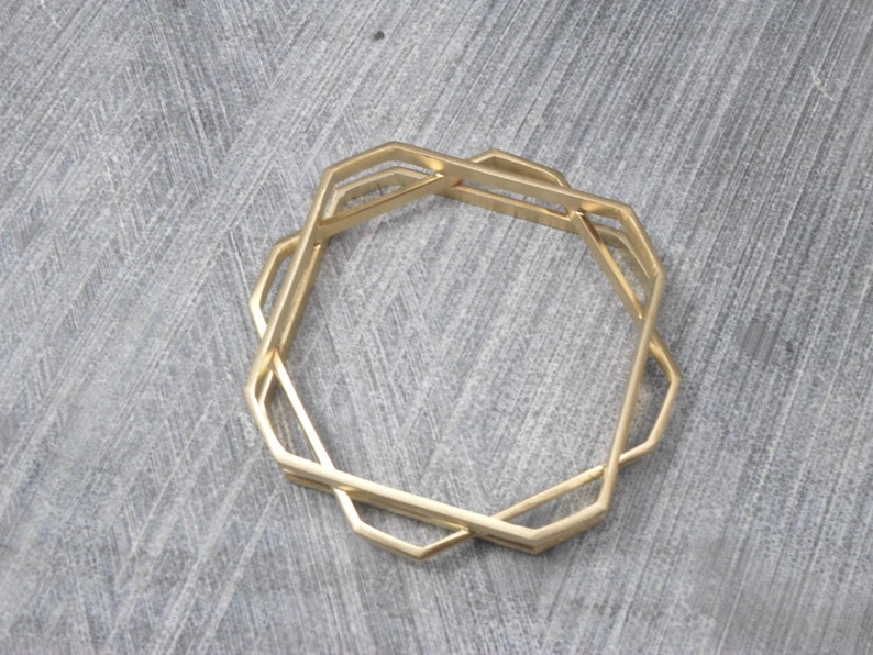 May include: A set of three gold-colored geometric bangles. Each bangle has a unique octagonal shape with interlocking designs. The bangles are displayed on a gray surface, showcasing their modern and minimalist style.