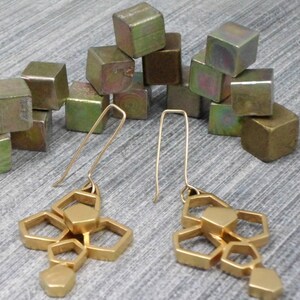 May include: Gold geometric earrings with a cluster of hexagonal shapes hanging from a thin gold wire. The earrings are displayed on a surface with a pile of small, metallic cubes.