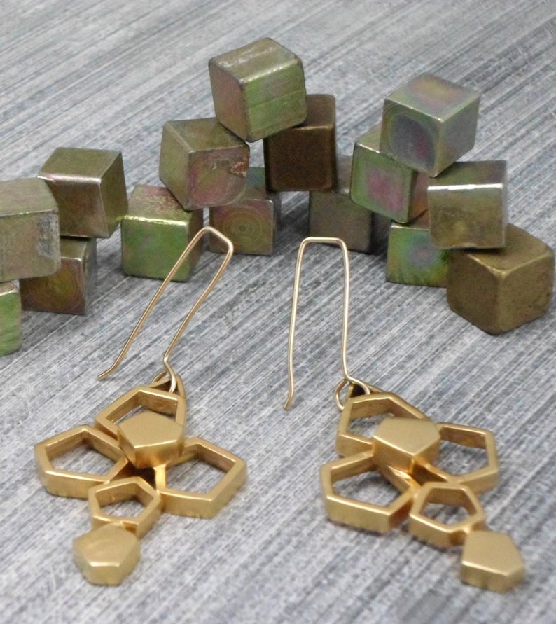 May include: Gold geometric earrings with a cluster of hexagonal shapes hanging from a thin gold wire. The earrings are displayed on a surface with a pile of small, metallic cubes.