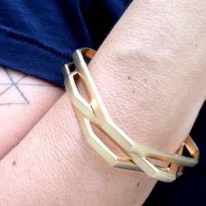 May include: A gold-colored geometric bracelet with a linked design. The bracelet is worn on the arm, with a dark blue shirt visible in the background. The bracelet's design features angular shapes and a polished finish.