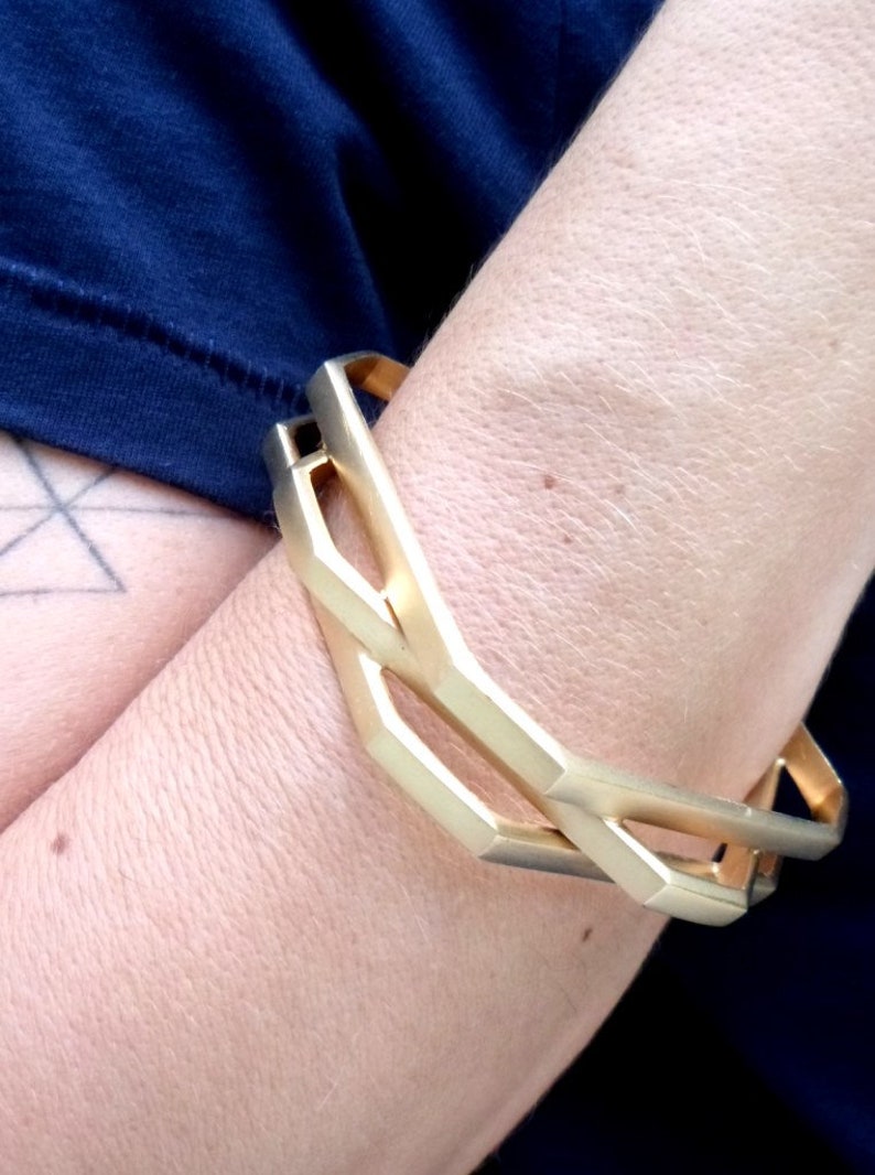 May include: A gold-colored geometric bracelet with a linked design. The bracelet is worn on the arm, with a dark blue shirt visible in the background. The bracelet's design features angular shapes and a polished finish.
