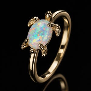 May include: A gold ring featuring a turtle-shaped design with a central oval-shaped iridescent opal stone. The turtle's shell is formed by the opal, while the head, four legs, and tail are crafted from polished gold metal with a smooth finish.