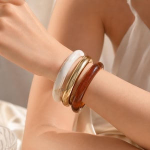 May include: A set of three stacked bangle bracelets worn on the wrist. The set includes one white marbled resin bangle, one polished gold-tone metal bangle, and one translucent amber-colored resin bangle, all featuring a smooth, rounded design.
