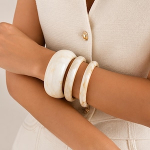 Op de afbeelding: Een set van drie gladde, crèmekleurige harsarmbanden met subtiele bruine marmering. De collectie bevat één brede, dikke manchetarmband en twee dunnere, ronde armbanden, allemaal voorzien van een gepolijste, glanzende afwerking en een neutrale tint.