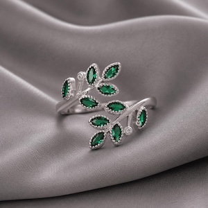Op de afbeelding: Een zilverkleurige open ring met twee vertakte stengels versierd met smaragdgroene marquise-geslepen stenen en kleine heldere accentstenen. Het ontwerp bootst een bladerige wijnstok na, waarbij de uiteinden van de ring elkaar licht overlappen.