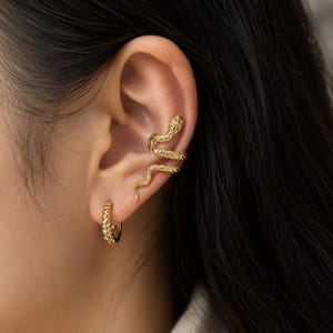 May include: A gold-toned ear cuff shaped like a coiled snake with a textured, scale-like surface, positioned along the upper cartilage, paired with a small, matching gold-toned hoop earring featuring a twisted rope design on the earlobe.