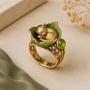 Puede incluir: Un anillo dorado que presenta un gato durmiendo enroscado sobre una hoja esmaltada en verde. La banda tiene un diseño calado intrincado similar a ramas, con una pequeña piedra redonda, translúcida y de color verde en el lateral.