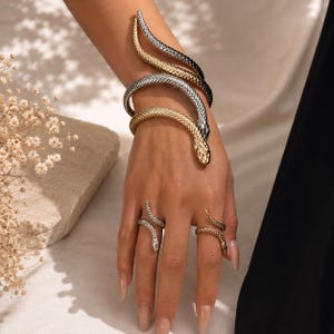 May include: A coiled snake arm cuff featuring a textured metallic finish in alternating gold and silver tones, accompanied by two matching snake rings worn on the fingers, each displaying a detailed scale pattern and a serpent head design.
