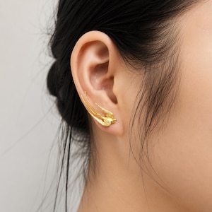 May include: A gold-toned ear climber earring shaped like a swallow in flight. The bird's body rests on the earlobe, while its long, slender wings extend upward along the outer edge of the ear cartilage, creating a sleek and continuous silhouette.