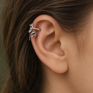 May include: A silver-toned ear cuff shaped like a small frog, featuring detailed limbs and a textured finish. The frog is positioned to appear as if it is clinging to the outer edge of the ear cartilage, with its front legs extended forward.