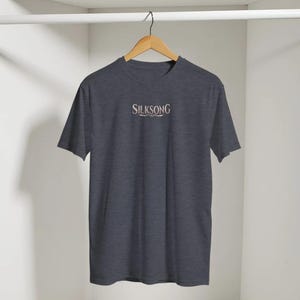May include: A dark heather grey short-sleeve t-shirt featuring the word SILKSONG printed in a light, decorative serif font across the centre of the chest. The garment is displayed on a wooden hanger against a plain, light-coloured background.
