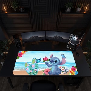 Op de afbeelding: Een rechthoekige bureaumat met een blauw alien-personage dat op een zandstrand zit met een turquoise oceaan op de achtergrond. Het ontwerp bevat kleurrijke tropische hibiscusbloemen, groene palmbladeren en blauwe vlinders op het oppervlak.