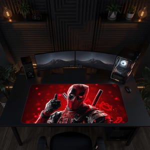 May include: A rectangular desk mat featuring a graphic illustration of the character Deadpool in a red and black suit, giving a thumbs-up gesture against a vibrant red background with bokeh light effects and two katana handles visible.
