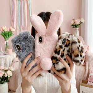 May include: Three plush phone cases featuring soft faux fur textures. One is solid grey, one is light pink with long rabbit ears and a round tail, and one features a brown and black leopard print. Each case has a decorative rhinestone camera bezel.