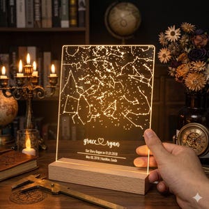 Peut inclure: Une veilleuse rectangulaire en acrylique avec une carte du ciel étoilé illuminée. La base est en bois clair. Le texte indique : grace heart ryan, Our Story Began on 01.01.2018, May 09, 2018 | Hamilton, Canda.