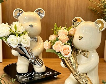 May include: Two glossy cream-colored ceramic bear figurines, each wearing a gold bow tie and holding a bouquet of artificial roses. One bear holds white roses, the other peach roses. The left bear stands on a black base with the text Mitra & Chat.