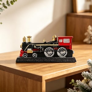 Pode incluir: Uma estatueta decorativa de locomotiva a vapor com corpo preto, detalhes vermelhos na frente e na cabine, acabamentos dourados na chaminé e na caldeira, e rodas prateadas, montada sobre uma base retangular preta com textura.