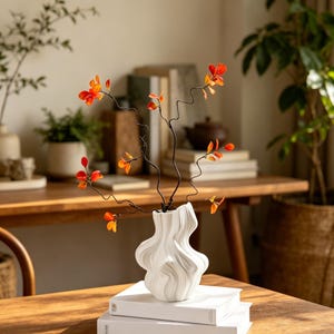 Könnte beinhalten: Eine weiße Keramikvase mit wellenförmiger, organischer Form enthält dünne, dunkle, gewundene Zweige, die mit kleinen, leuchtend orangefarbenen Blättern geschmückt sind. Die Vase steht auf zwei gestapelten weißen Büchern auf einer Holzoberfläche.