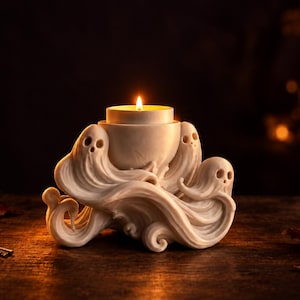 Peut inclure: Un bougeoir de couleur crème sculpté avec plusieurs silhouettes de fantômes tourbillonnant autour d'une ouverture circulaire centrale. Une petite bougie chauffe-plat allumée est placée à l'intérieur, diffusant une lueur chaude.