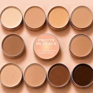 May include: Nine round metal tins containing cream tallow foundation in various skin tones. The center tin features a peach label with the text PRETTY IN PEACE, cream tallow foundation, hydrating + blurring, blendable + long-wear, net wt. 1.20 oz.