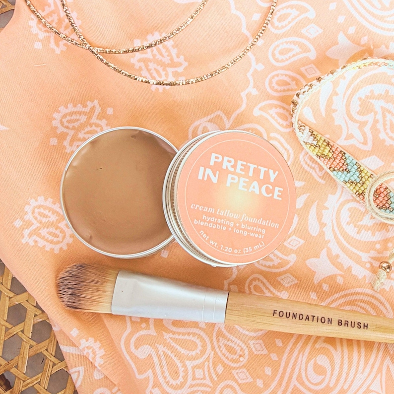 May include: A round metal tin of Pretty In Peace cream tallow foundation with a net weight of 1.20 oz, featuring a peach-colored label with white text. Beside the open tin lies a makeup brush with a light wood handle labeled Foundation Brush.