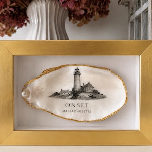 May include: A rectangular gold frame containing a white oyster shell with a gold-painted edge. The shell features a black and white illustration of a lighthouse and the text ONSET MASSACHUSETTS. A small white pearl is attached to the left side.