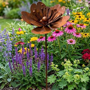 May include: A rust-colored metal flower sculpture featuring layered petals and a central stamen with spherical tips, mounted on a thin vertical metal rod. The sculpture is positioned among various garden flowers including purple salvia and pink coneflowers.