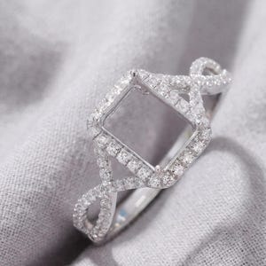 Op de afbeelding: Een zilverkleurige ringzetting met een vierkante centrale houder voor een edelsteen. De band en het vierkante frame zijn ingelegd met talloze kleine, heldere, fonkelende steentjes en hebben een gespleten, gedraaid ontwerp.