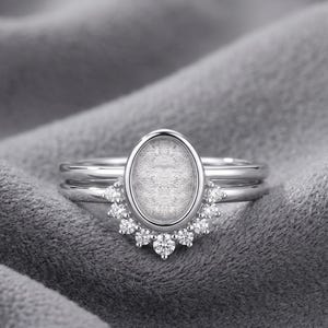 Könnte beinhalten: Ein silberfarbener Ring mit einer zentralen ovalen Fassung und einer strukturierten grauen Einlage. Die Basis des Ovals ist mit einer halbkreisförmigen Anordnung aus neun kleinen, klaren, rund geschliffenen Steinen verziert.