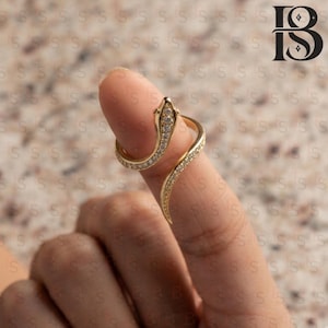 May include: A gold-toned snake ring featuring a coiled design that wraps around the finger. The surface of the snake is encrusted with small, clear, sparkling stones along its entire length, including the head and the tapering tail section.