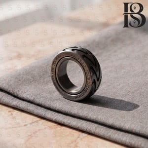 Puede incluir: Un anillo de metal gris oscuro que presenta un patrón de banda de rodadura de neumático alrededor de la banda exterior. El anillo está grabado con el texto "VERAFIE TOCH" y "FEED YOUR INSPIRATION" a lo largo de los bordes lisos de la banda.