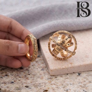 May include: A gold-toned metal ring featuring intricate engraved patterns on its outer surface, displayed alongside a spherical armillary ring that unfolds into a band, both pieces decorated with etched lettering and symbols in a metallic finish.