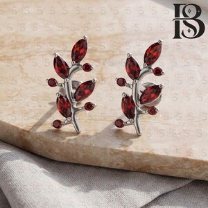 May include: A pair of silver-toned stud earrings shaped like branches, each featuring multiple marquise-cut and round-cut deep red garnet gemstones arranged along the metal stems. The earrings have a polished finish and a secure post-back design.