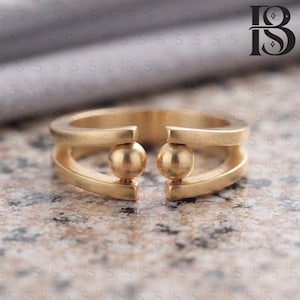 May include: A gold-toned open ring featuring two parallel bands connected by two small, spherical beads positioned at the center gap. The ring has a smooth, polished finish and a wide, structured design that remains open at the front.
