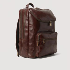 Puede incluir: Una mochila de cuero marrón oscuro que presenta un compartimento principal grande con cremallera superior, un bolsillo frontal secundario con cremallera y un asa superior. La bolsa incluye múltiples cremalleras metálicas con tiradores.
