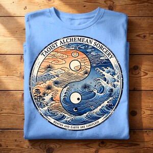 May include: A light blue t-shirt featuring a circular graphic of a yin-yang symbol composed of stylised waves and clouds. The text TAOIST ALCHEMEAN SORCERY is printed along the top edge, and HEAVEN AND EARTH ARE LIMITLESS is printed along the bottom edge.
