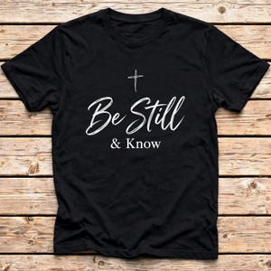May include: A black short-sleeved t-shirt featuring a white graphic print on the chest. The design includes a small cross symbol positioned above the white text that reads Be Still & Know in a combination of script and sans-serif typography.