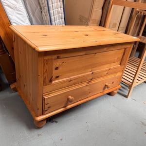 May include: A rectangular wooden storage chest with a natural light brown finish and visible wood grain. The chest features a flat top surface, a single horizontal panel on the upper front, and one lower drawer equipped with two small round wooden knobs.