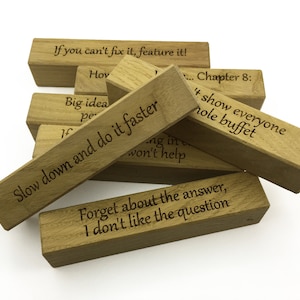 May include: Wooden blocks with humorous sayings engraved on them. The sayings include "Slow down and do it faster", "If you can't fix it, feature it!" and "Forget about the answer, I don't like the question".
