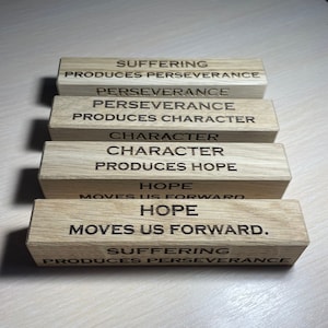 May include: A set of six wooden blocks with inspirational quotes engraved on each side. The quotes are: "Suffering produces perseverance", "Perseverance produces character", "Character produces hope", "Hope moves us forward", "Suffering produces perseverance".