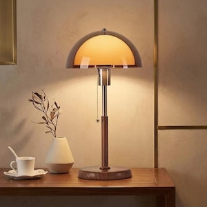 May include: A table lamp featuring a dome-shaped amber glass shade, a brushed metal neck, and a dark wood base and stem. The lamp includes a pull chain switch and emits a warm, soft glow. The design combines glass, metal, and wood elements.